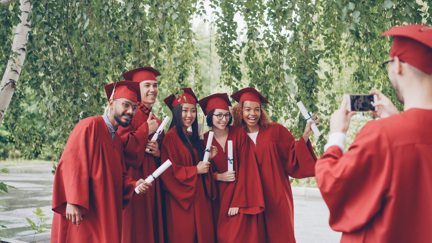 Lima wisudawan berjas merah tersenyum memegang ijazah di luar ruangan.