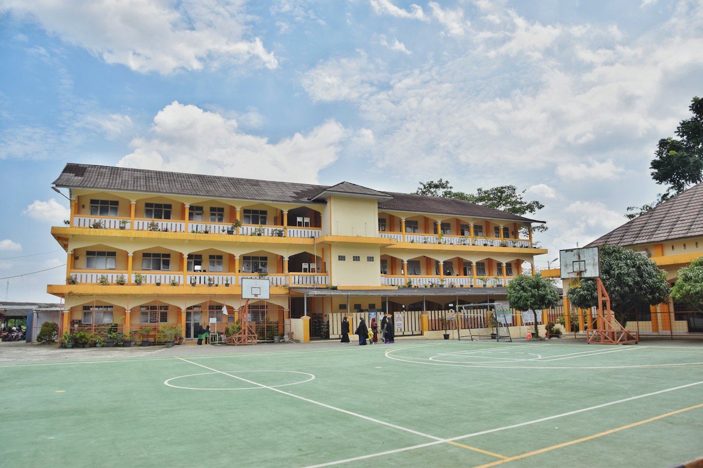 Orang-orang berjalan di dekat gedung sekolah berwarna kuning dengan lapangan basket.