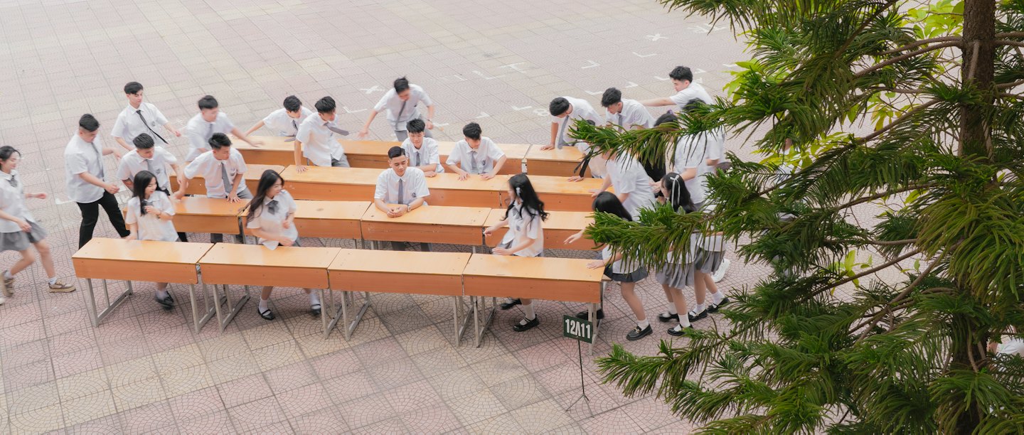 Siswa berseragam bersosialisasi di sekitar meja kayu di halaman sekolah.